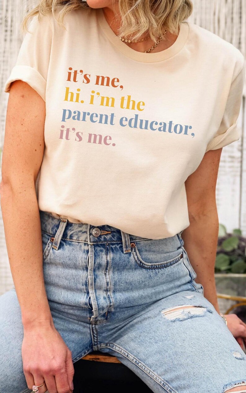 Pu&ograve; includere: Una maglietta color crema con la scritta "it's me, hi. I'm the parent educator, it's me." in lettere colorate. La maglietta &egrave; abbinata a jeans di denim blu chiaro con dettagli strappati.