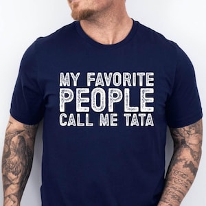 May include: Navy blue t-shirt with the white text "MY FAVORITE PEOPLE CALL ME TATA". The shirt is a crew neck style and appears to be made of a soft material. The text is in a distressed font.