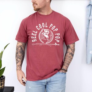 May include: A red t-shirt with a white graphic of a fish with the text "Reel Cool Pop Pop" around it.