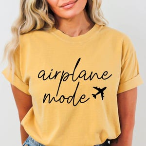 Op de afbeelding: Een geel T-shirt met zwarte tekst die "vliegtuigmodus" zegt en een kleine vliegtuigafbeelding.