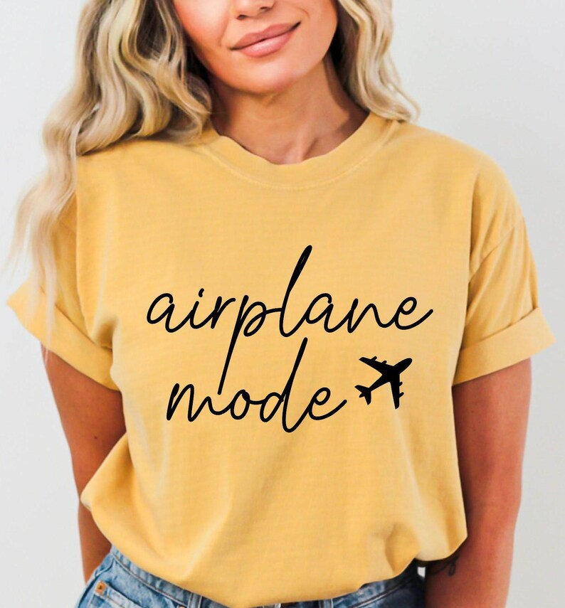 Op de afbeelding: Een geel T-shirt met zwarte tekst die "vliegtuigmodus" zegt en een kleine vliegtuigafbeelding.