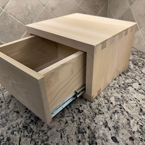 May include: A light-colored wooden drawer with dovetail joints, partially open to reveal the interior. The drawer sits on a granite countertop. The drawer slides are visible. The overall design is simple and modern.