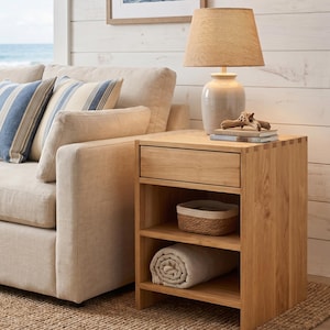 May include: A light wood nightstand with a drawer and two open shelves. A woven basket and rolled blanket are on the shelves. A beige lamp with a woven shade sits on top, next to a stack of books and driftwood.