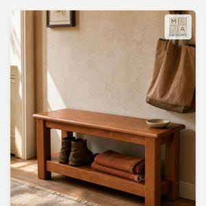 May include: A rectangular wooden bench with a smooth top surface and a lower shelf. The bench features four sturdy legs. On the lower shelf, there is a pair of brown leather boots and a folded orange textile. A small ceramic bowl sits on the bench top.