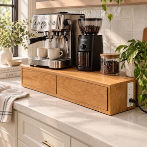 May include: A rectangular wooden coffee station organizer with two front-facing drawers and a built-in white power strip on the right side. The organizer supports a stainless steel espresso machine, a black coffee grinder, and a glass jar of beans.