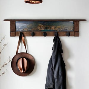 May include: A wooden wall-mounted coat rack with a landscape print and five black hooks. A brown hat with a copper-colored interior hangs from the rack, along with a black jacket. The rack is mounted on a white wall.
