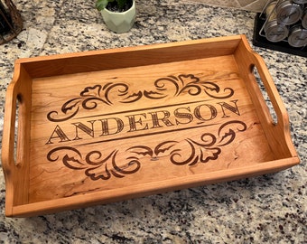 May include: A wooden serving tray with a natural wood finish. The tray has two handles and is engraved with the name "ANDERSON" in a decorative font with a floral design.