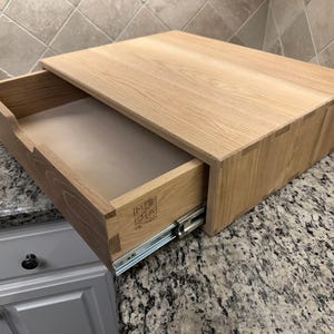 May include: A wooden drawer with dovetail joints, partially open, revealing the interior. The drawer is made of light-colored wood and sits on a countertop. The drawer's front has a logo. The drawer is designed for storage.