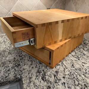 Peut inclure: Deux tiroirs en bois, dont un partiellement ouvert, empilés l'un sur l'autre. Les tiroirs sont en bois clair et présentent des assemblages à queue d'aronde. Le tiroir ouvert révèle le logo "MEJA DESIGN".