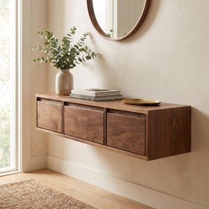 May include: A floating wooden console with three drawers, showcasing a warm, natural wood grain. A round mirror hangs above, reflecting the room. A vase with greenery, books, and a small gold tray adorn the top. The console is mounted on a neutral-colored wall.