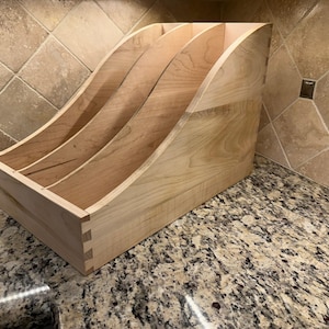 May include: A wooden magazine rack with three compartments. The rack is made of light wood and has a curved front.
