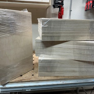 May include: A stack of four sheets of plywood wrapped in plastic, with one sheet standing upright next to the stack.