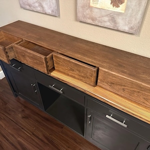 Puede incluir: Una consola de madera oscura con tres cajones abiertos. La mesa tiene una base negra con tiradores plateados y una tapa de madera a rayas. Los cajones están abiertos, revelando la construcción interior. La mesa está contra una pared beige.