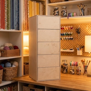 May include: A tall, light-colored wooden cabinet with five drawers stands on a wooden countertop. The cabinet is in a craft room with shelves filled with fabric rolls, baskets of yarn, and jars of sewing supplies.