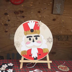 Puede incluir: Decoración navideña circular de madera con un diseño de cascanueces. El cascanueces tiene un sombrero y una chaqueta rojos, una barba blanca y detalles dorados. La decoración se exhibe en un pequeño caballete de madera, con recortes de copos de nieve.