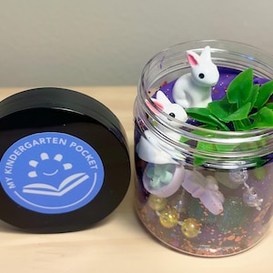 Peut inclure: Un pot en plastique transparent rempli de slime violet, de paillettes et de petits jouets, dont deux figurines de lapins blancs et des plantes en plastique vertes. Le pot a un couvercle noir avec un autocollant bleu qui dit "My Kindergarten Pocket".