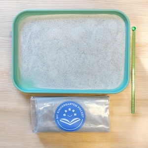 May include: A light blue plastic tray filled with white sand. A clear plastic bag with a blue logo that says "My Kindergarten Pocket" is next to the tray. A green plastic ruler is on the right side of the image.