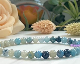Puede incluir: Una pulsera de cuentas redondas en tonos azul claro, blanco y azul oscuro. Las cuentas están ensartadas. La palabra "Aquatice" está escrita en rosa. El fondo presenta un arreglo floral borroso.