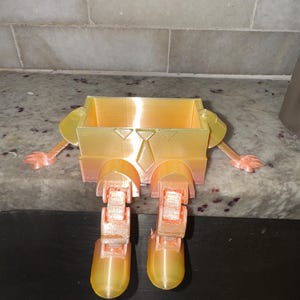 May include: A 3D-printed, box-shaped planter with arms and legs. The planter is primarily yellow with orange accents, including the arms, legs, and hands. The design resembles a cartoon character, with a tie detail on the front.