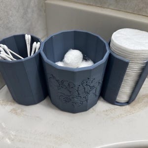 May include: A set of three matte slate blue bathroom organizers with a faceted, geometric exterior. The left container holds white cotton swabs, the center container holds white cotton balls, and the right container holds a stack of white cotton pads.