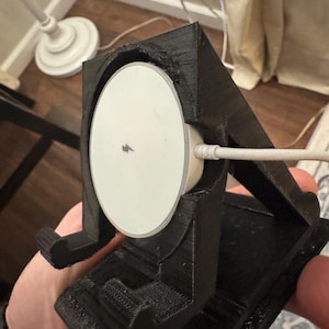 May include: A black 3D-printed charging stand for a wireless charger. The stand holds a white charging pad with a charging cable attached. The stand is held in a person's hand.