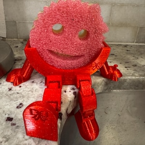 May include: A red 3D-printed sponge holder shaped like a cartoon character, with arms, legs, and a smiling face. The sponge is pink and fits into the holder. The holder is sitting on the edge of a sink.