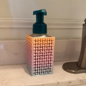 May include: A 3D-printed soap dispenser with a woven basket design. The dispenser's body transitions from orange to pink to blue. It has a dark teal pump and sits on a white countertop. The dispenser is a bathroom accessory.