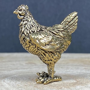 May include: A small, metallic gold-coloured chicken figurine featuring detailed, textured feather patterns across its body, a defined comb on its head, and a sharp beak, standing in a profile position with its head turned slightly towards the left.