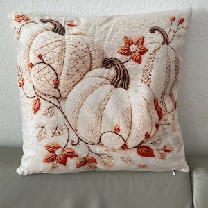 Könnte beinhalten: Ein quadratisches Zierkissen mit einem cremeweißen Stoffbezug und gestepptem Muster. Das Design zeigt drei gestickte Kürbisse in Creme- und Beigetönen, akzentuiert durch braune Stiele und leuchtend orangefarbene Herbstblätter.