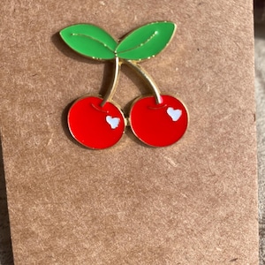 Puede incluir: Un broche de esmalte y metal dorado con forma de dos cerezas con frutos de color rojo brillante, hojas verdes y pequeños detalles blancos en forma de corazón en cada cereza. El broche tiene un acabado liso y brillante con bordes dorados.