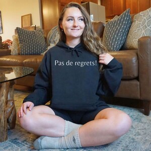 May include: A woman wearing a black hoodie with the text "Pas de regrets!" in white. She is sitting on a rug with her legs crossed.