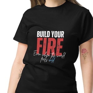 Könnte beinhalten: Schwarzes T-Shirt mit dem Text "BUILD YOUR FIRE" in Weiß und Rot sowie dem Satz "Even when the world feels cold" in Schreibschrift. Das Model trägt eine blaue Jeans.