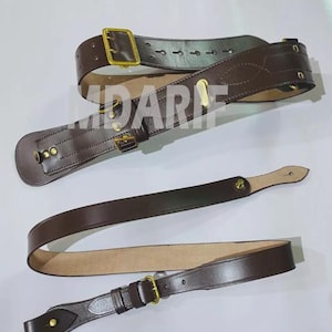 May include: A brown leather belt with a brass buckle and a strap. The belt is designed for carrying a weapon or other equipment.
