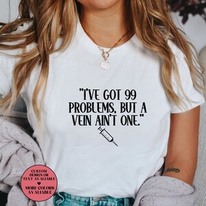 May include: White t-shirt with black text that reads "I've got 99 problems, but a vein ain't one." A black and white syringe is below the text. The shirt has a pink circle with white text that reads "Custom design or text available" and a pink heart. Below the heart is white text that reads "More colours available".