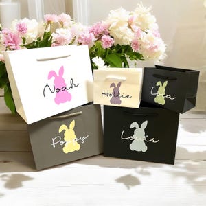 May include: Four gift bags with bunny silhouettes and names printed on them. The bags are white, grey, black, and beige. The bunnies are pink, yellow, green, and purple.