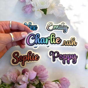 May include: A selection of name stickers with a holographic finish. The names "Alex", "Emily", "Charlie", "Sarah", "Sophie", and "Poppy" are shown in different colours and fonts. The stickers are surrounded by pink and white flowers.