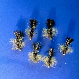 May include: Six fly fishing lures with brown, black, and white feathers and iridescent accents on a blue background.