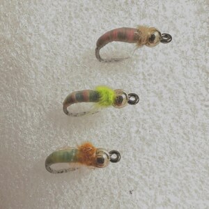 May include: Three fly fishing lures with different colored bodies and bead heads. The lures are on a white background.