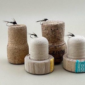 May include: Four cork bottle stoppers with fly fishing lures attached. The stoppers are made of wood and cork and have different colors and patterns. The lures are black and silver and have feathers.