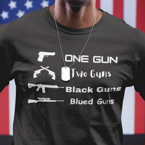 May include: A black t-shirt with a white graphic of different types of guns and the text "One Gun", "Two Guns", "Black Guns", and "Blued Guns".