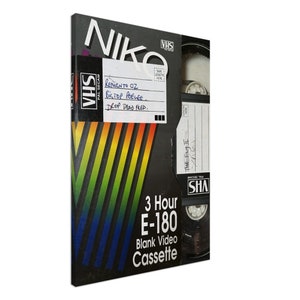 Könnte beinhalten: Eine schwarze VHS-Kassette mit einem Etikett, auf dem "3 Hour E-180 Blank Video Cassette" steht. Das Etikett hat ein Regenbogenstreifen-Design. Das Band ist mit dem Text "Return to OZ Bictop PeeLee. Drop Dead Fred." beschriftet.