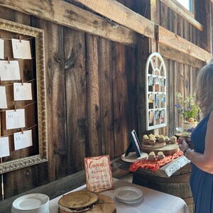 Puede incluir: Una rústica decoración de recepción de boda con un fondo de madera. Una mesa exhibe cupcakes, platos y un letrero que dice "Cupcake Flavors". Un plano de asientos enmarcado cuelga a la izquierda.