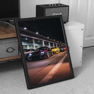 May include: Framed print showing a line-up of sports cars at night. The cars are various colours, including red, yellow, and silver, parked on a lit road. The image is in a black frame, resting on a carpeted surface.