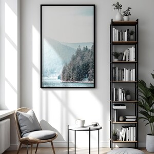 Pode incluir: Um interior de sala de estar com uma fotografia emoldurada em preto de uma paisagem montanhosa nevada, uma cadeira de vime com um almofada cinza, uma mesa auxiliar preta com um tampo branco, uma caneca branca, um pufe cinza e uma estante de metal preto com prateleiras de madeira cheias de livros e plantas.