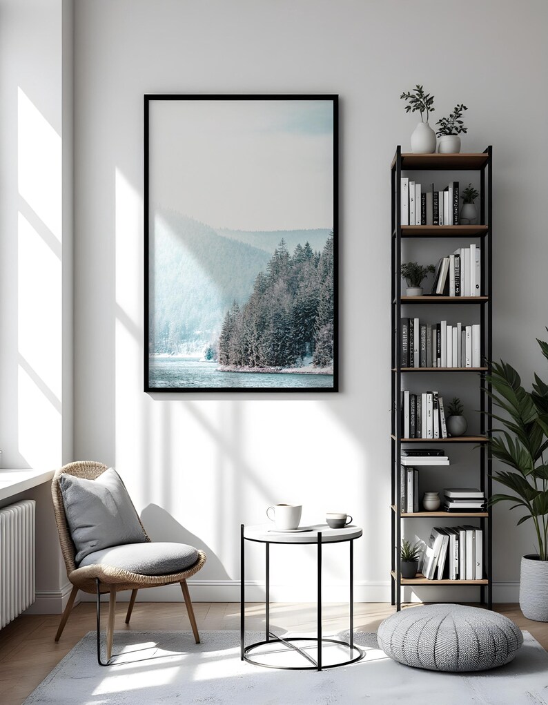 Pode incluir: Um interior de sala de estar com uma fotografia emoldurada em preto de uma paisagem montanhosa nevada, uma cadeira de vime com um almofada cinza, uma mesa auxiliar preta com um tampo branco, uma caneca branca, um pufe cinza e uma estante de metal preto com prateleiras de madeira cheias de livros e plantas.