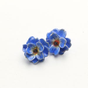 Op de afbeelding: Twee blauwe bloemvormige oorbellen met een donker centrum en gouden rand. Ze zijn geplaatst tegen een witte achtergrond. Het bloemendesign is gedetailleerd en realistisch, wat wijst op een door de natuur geïnspireerd thema.