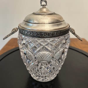 May include: A clear glass jar with a diamond-cut pattern and a silver-toned metal lid. The lid features a decorative finial handle at the top and two ornate metal side handles attached to the rim. The glass body has a tapered, rounded shape.