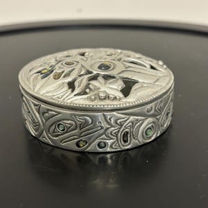 May include: A silver-coloured, round trinket box with intricate, carved designs. The lid features openwork floral and geometric patterns, with inlaid iridescent accents. The sides of the box are adorned with detailed, stylized artwork. The box is on a dark surface.