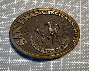 Puede incluir: Hebilla de cinturón ovalada de color bronce con letras e imágenes en relieve. El texto dice "SAN FRANCISCO MUSEUM OF ART" y "SOAP BOX DERBY". El diseño central presenta un carro tirado por caballos con un conductor.