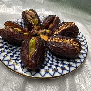 May include: A plate of Medjool dates, some stuffed with pistachios and others with sesame seeds. The dates are arranged on a white and blue patterned plate with a gold rim. The background is a white, crinkled surface.
